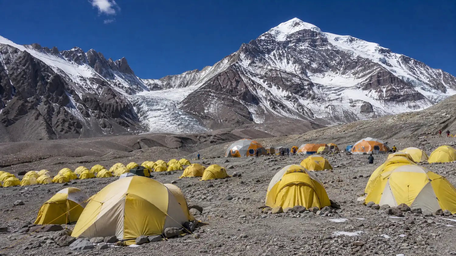 Reihen gelber und orangefarbener Expeditionszelte auf einer felsigen Moräne im Basislager Plaza de Mulas, dahinter verschneite Gipfel und ein Gletscher