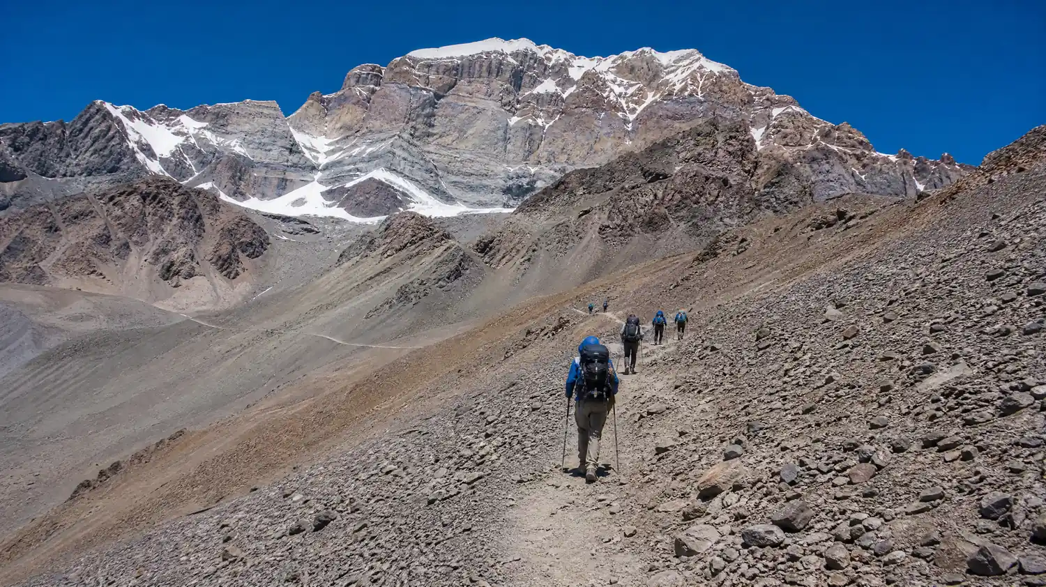 Wanderer von hinten auf einem langen Zickzackpfad durch Geröll in großer Höhe, trockene braune und graue Berge mit Schneefeldern