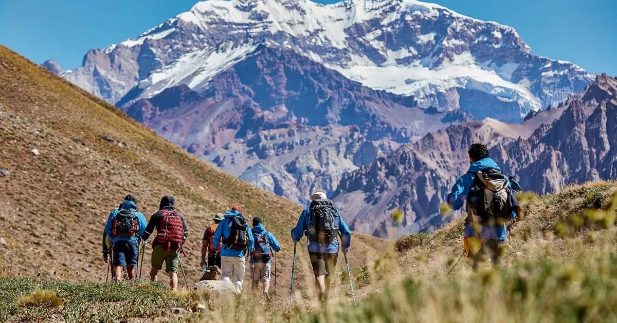 Eine Reihe von Wanderern mit Tagesrucksäcken auf dem Weg durch ein grasbewachsenes Tal hinauf zu einem verschneiten Berg