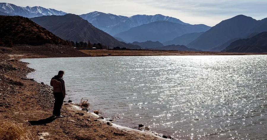 Ein Wanderer steht an einem steinigen Ufer über einem sonnenbeschienenen Bergsee in den Andenvorbergen