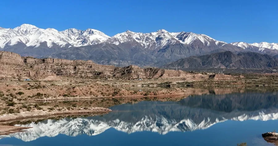 Schneebedeckte Anden spiegeln sich im stillen Stausee von Potrerillos unter rötlichen Felswänden