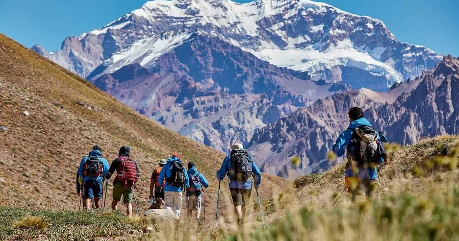 Eine Reihe von Wanderern mit Tagesrucksäcken geht ein grasiges Tal hinauf in Richtung des verschneiten Aconcagua